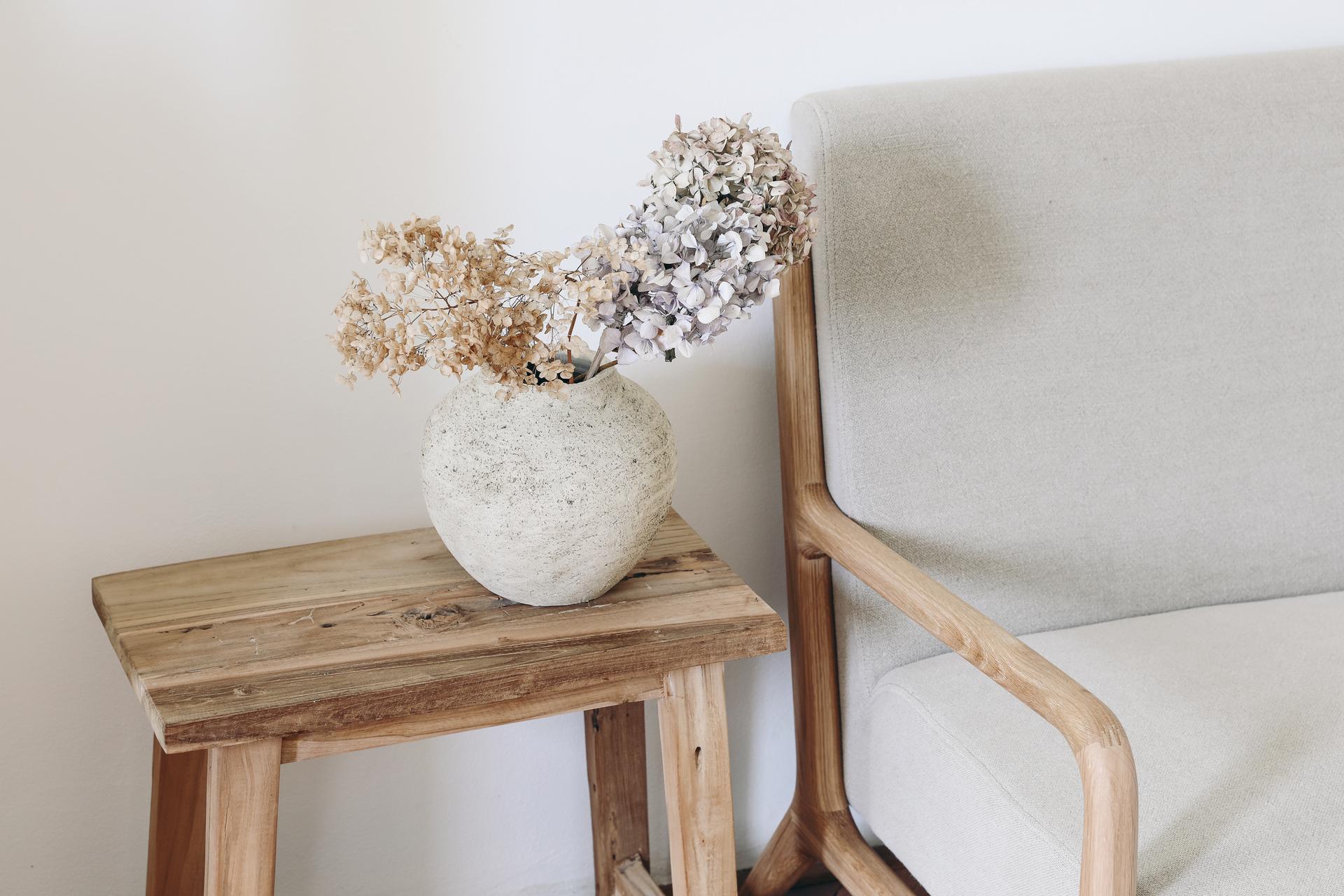 Fall still life photo. Rustic textured vase with dry hydrangea flowers on old wooden bench. Blurred linen mid century sofa background. White wall. Scandinavian interior. Boho elegant home decor.